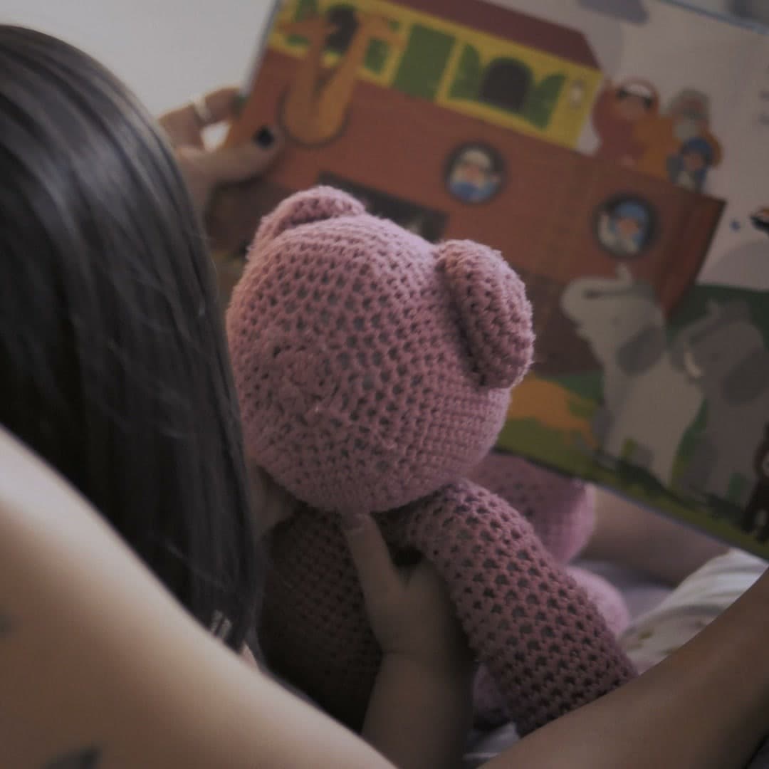 A child holding a knitted bear while an adult reads a picture book, seen from behind.